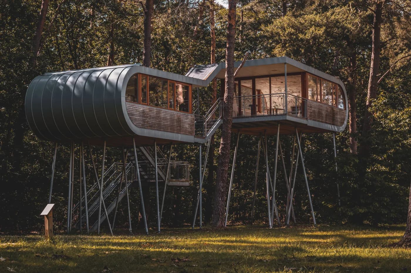 Imagen de ejemplo de BookNoFees: dos modernas casas en los árboles sobre pilotes, con escaleras metálicas entre los árboles del bosque