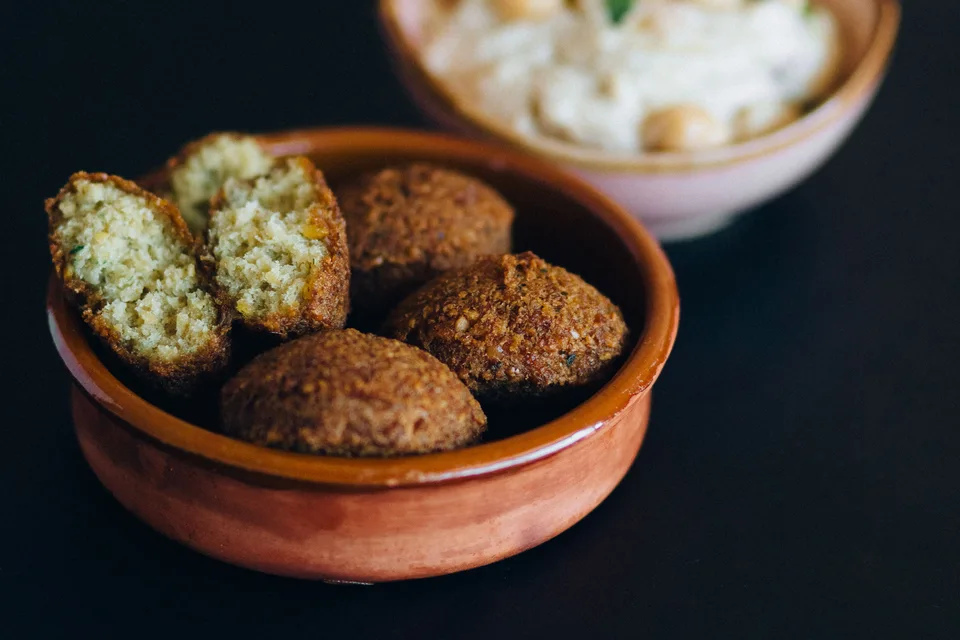 BookNoFees: Foto de ejemplo: Bolas de falafel, incluida una cortada por la mitad, en un cuenco con hummus detrás.